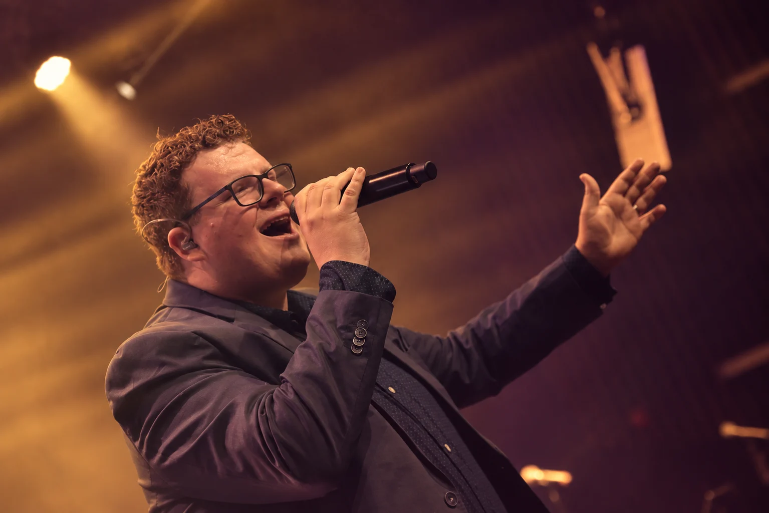 Stefan van Eijk met microfoon en uitgestrekte arm, in een warme kleurvariant van zijn podiumfoto